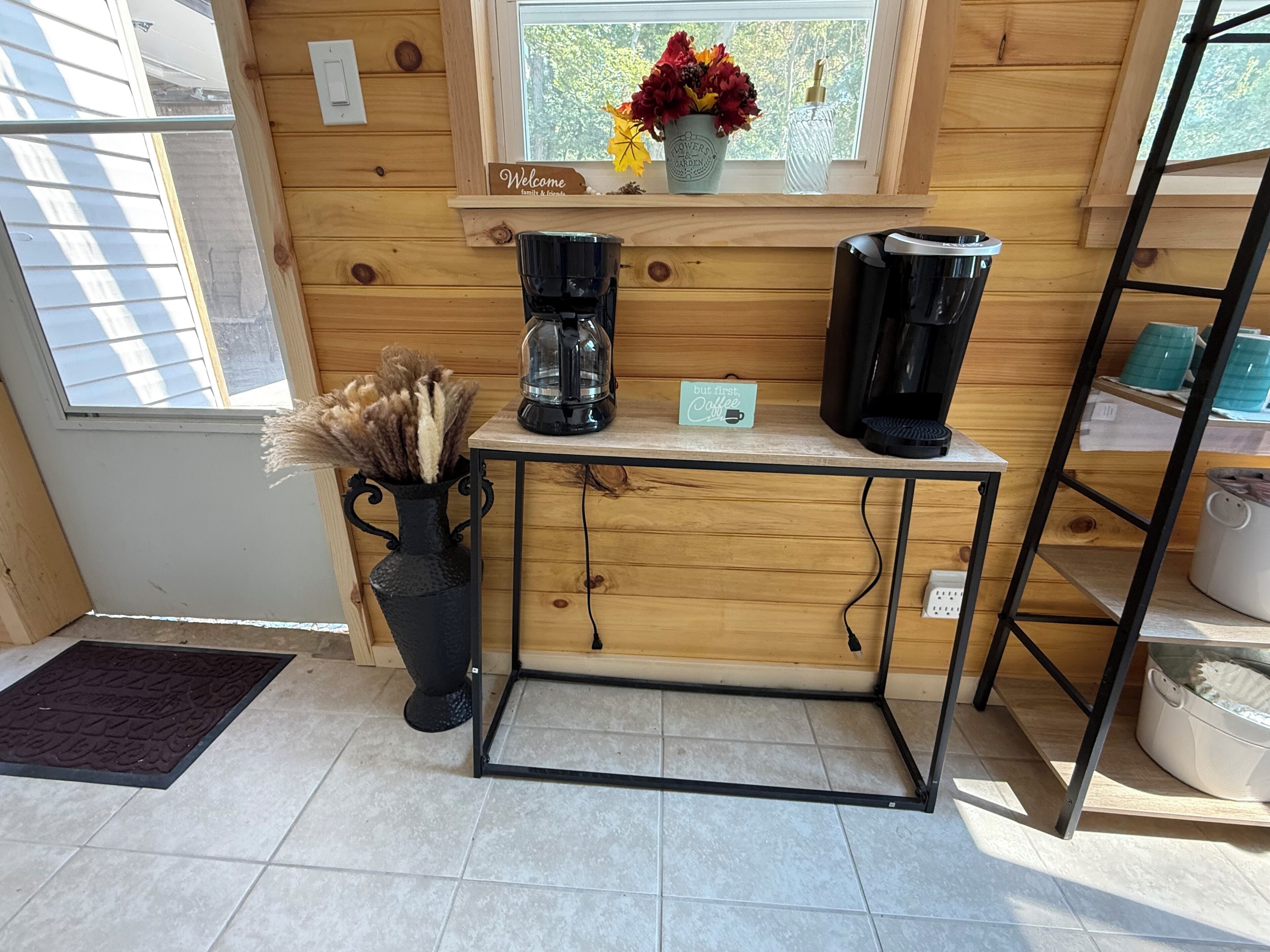 Un intérieur en bois avec un coin café comprenant deux machines à café, un panneau d’accueil et un vase avec des plantes séchées.