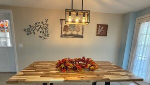 Een eettafel met een houten blad en zwarte metalen poten, een decoratieve lamp erboven en een bord met herfstbladeren aan de muur.