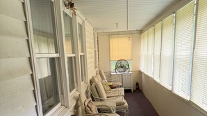 Un espacio de estar al aire libre cubierto, con sillas acolchadas y una mesa, un ventilador y una ventana con persianas.