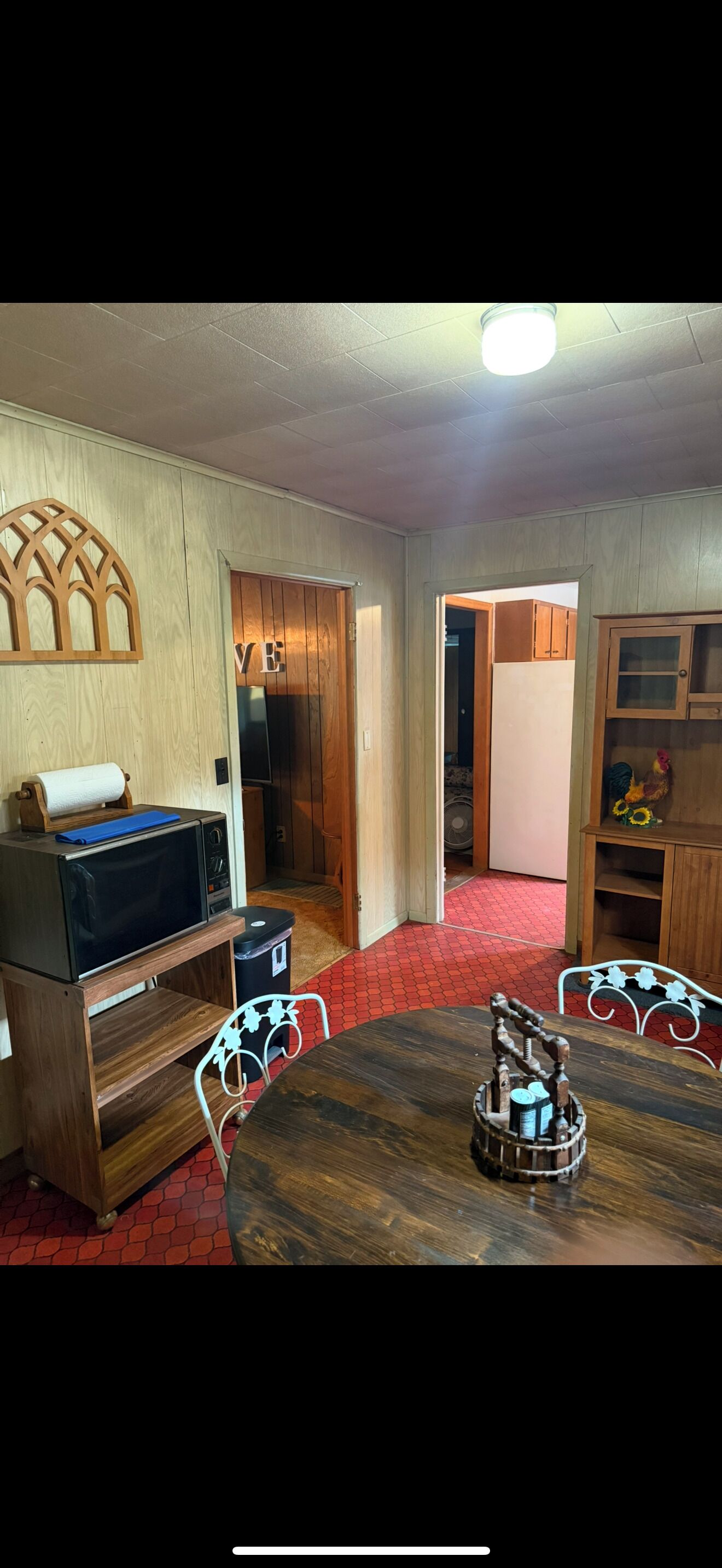 A room with a wooden table, a microwave on a wooden stand, a red patterned floor, and a wooden cabinet with a glass door.