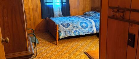 Une chambre avec des murs en bois, un lit recouvert d’une courtepointe à motifs, une armoire en bois et une petite table.