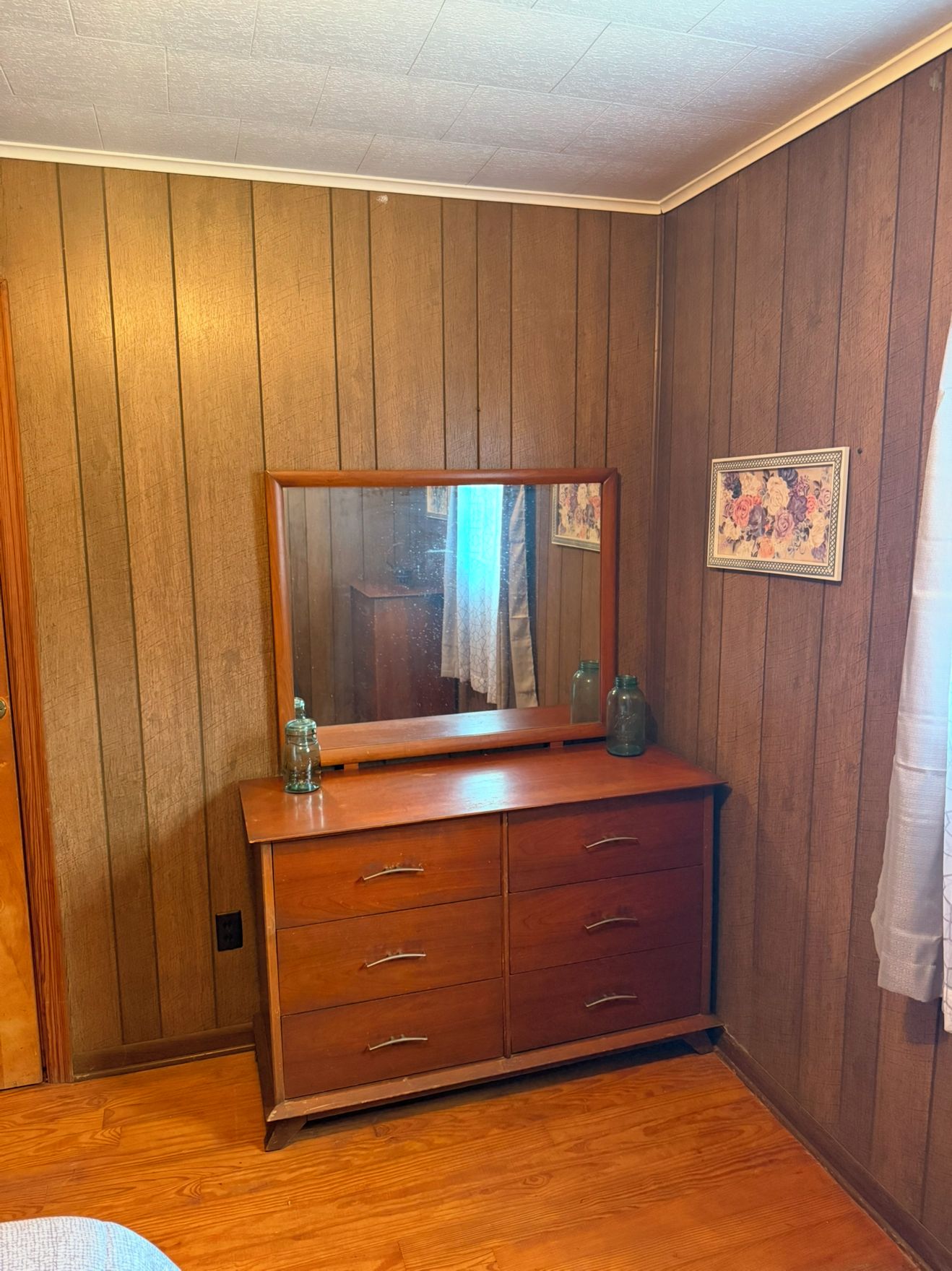 A room with wooden panel walls, a wooden dresser with drawers, a mirror, and a framed picture.