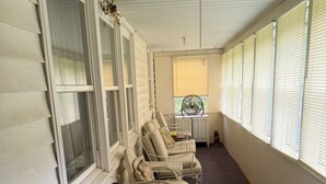 Un espace extérieur abrité, avec des chaises rembourrées et une table, un ventilateur et une fenêtre équipée de stores.