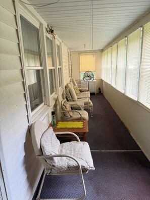 Un espace extérieur abrité, avec des chaises rembourrées et une table, un ventilateur et une fenêtre équipée de stores.