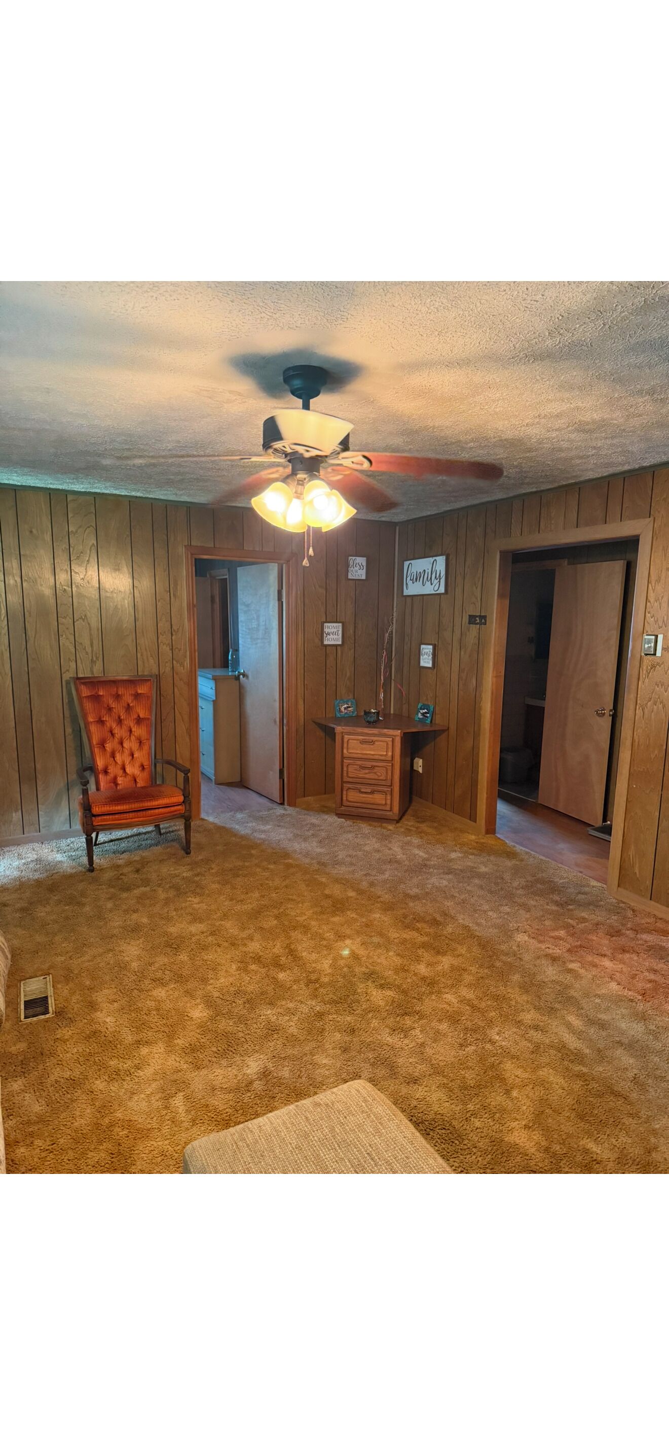 A room with wooden paneling, a ceiling fan, a red upholstered chair, a wooden cabinet, and a carpeted floor.
