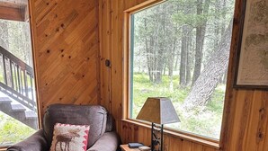 A cozy room with a wooden interior, a cushioned armchair, a small table with a lamp, and a large window offering a view of a forest.
