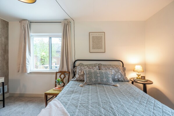A bedroom with a bed, a bedside table with a lamp, a chair, a window with curtains, and a framed picture on the wall.