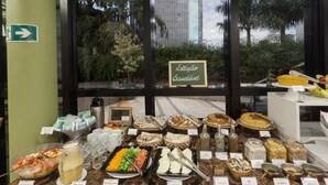 Un bufé con diversas opciones de comida y bebidas, un letrero que dice "Estação Saudavel", y una vista de palmeras y un edificio alto a través de la ventana.
