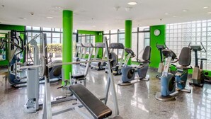 Un gimnasio con diversos equipos de ejercicio, ventanas amplias y pilares verdes.