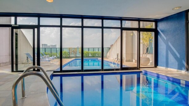 Una piscina interior con paredes de vidrio transparente, una escalera metálica y vistas a la ciudad.