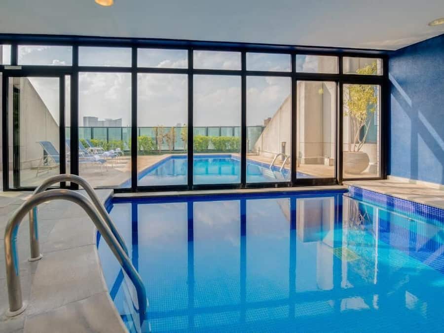 An indoor swimming pool with clear glass walls, a metal ladder, and a view of the cityscape outside.