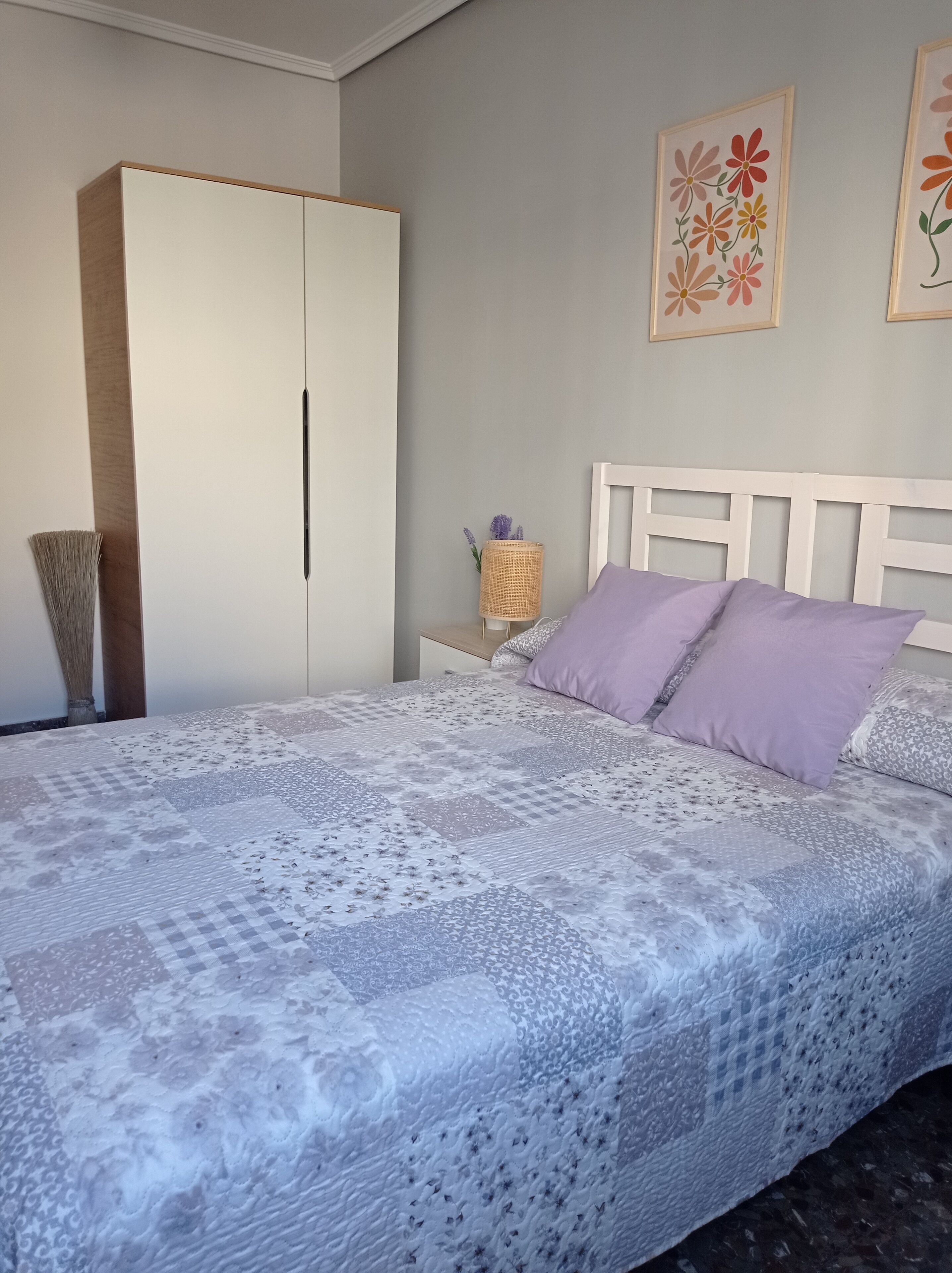 Un dormitorio con una cama grande, un armario, una mesita de noche con lámpara, y dos cuadros con motivos florales en la pared.