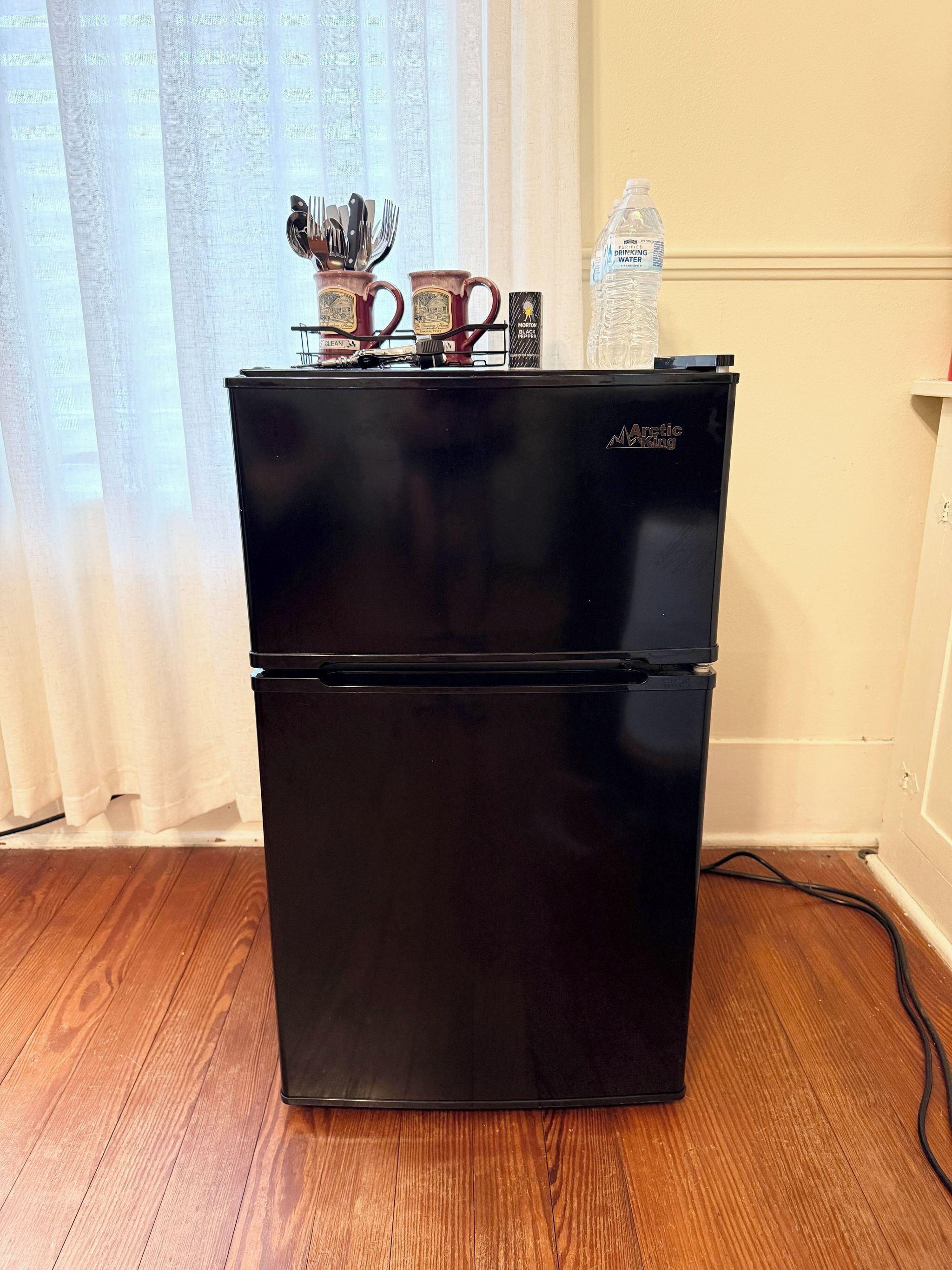 Un réfrigérateur noir de la marque Arctic King, doté d’un distributeur d’eau, avec une tasse et des ustensiles posés sur le dessus.