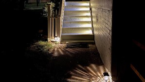 Un escalier menant à l’entrée d’un immeuble, de nuit, avec un éclairage extérieur qui illumine les marches et les environs.
