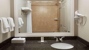Une salle de bain avec un grand miroir, un lavabo, une douche et un sèche-cheveux posé sur une étagère fixée au mur.