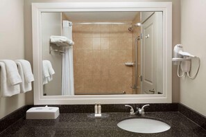 Un baño con un espejo grande, un lavamanos, una ducha y un secador de pelo sobre una estantería de pared.