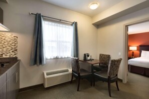 A hotel room with a dining table, two chairs, a bed, a nightstand, a lamp, and a window with curtains. - Travelodge by Wyndham Kindersley (Kindersley)