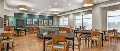 A spacious dining area with multiple tables and chairs, a green wall with framed pictures, and large windows allowing natural light.