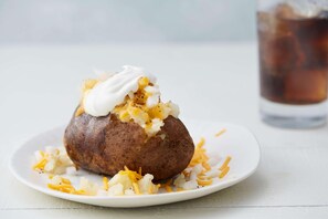 Bagt kartoffel med cremefraiche og revet ost, serveret på en hvid tallerken med et glas iste i baggrunden.