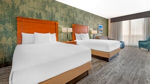 Une chambre d’hôtel avec deux lits, une grande fenêtre avec des rideaux, un mur décoré sur le thème d’une forêt verdoyante et une chaise bleue.