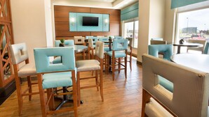 Un hall de hotel con suelo de madera, sillas tapizadas en azul claro, un televisor de pantalla plana montado en la pared y grandes ventanales con vistas al estacionamiento.