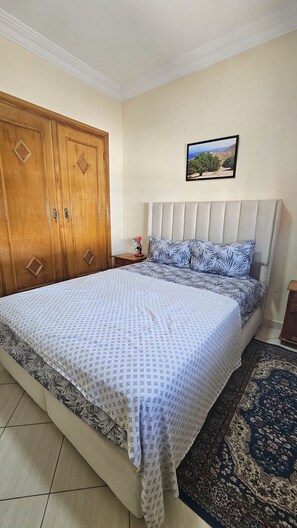Un dormitorio con cama, armario de madera, mesita de noche, un cuadro enmarcado en la pared y una alfombra estampada.