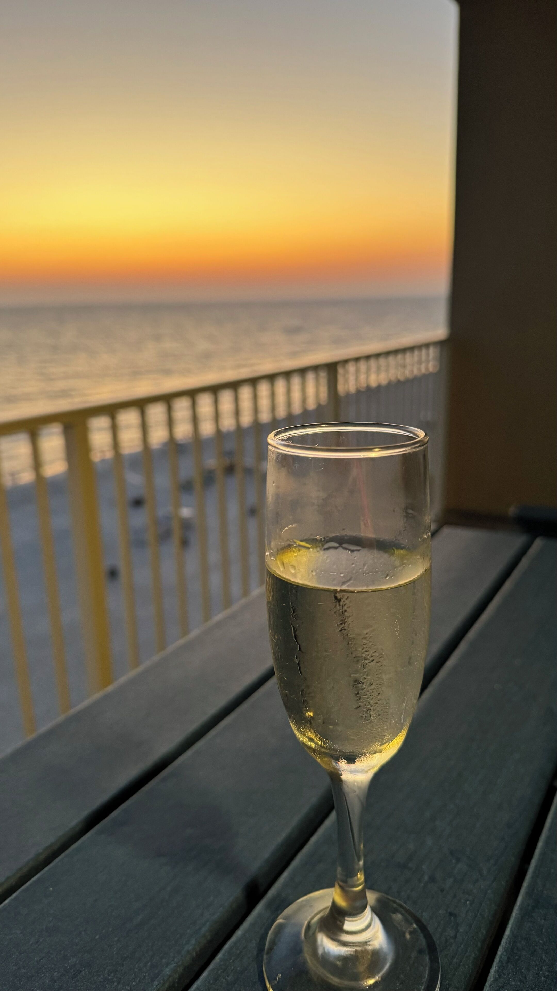 Una copa de vino blanco sobre una mesa de madera, con vista al océano y una puesta de sol al fondo.