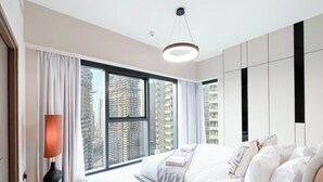 A modern bedroom with a large bed, a bedside table, a lamp, and a view of tall buildings through large windows. - Skyline Elegance Fountain View 3BR in Downtown (Dubai)
