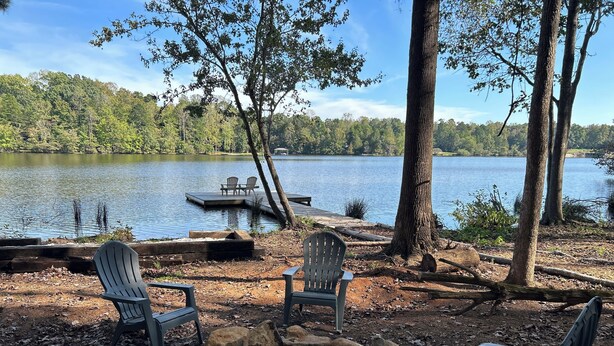 A lakeside seating area with Adirondack chairs and a fire pit, surrounded by trees and a dock extending into the water.