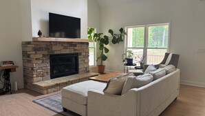 A living room with a stone fireplace, a flat-screen TV, a large window, a sofa, and a potted plant.