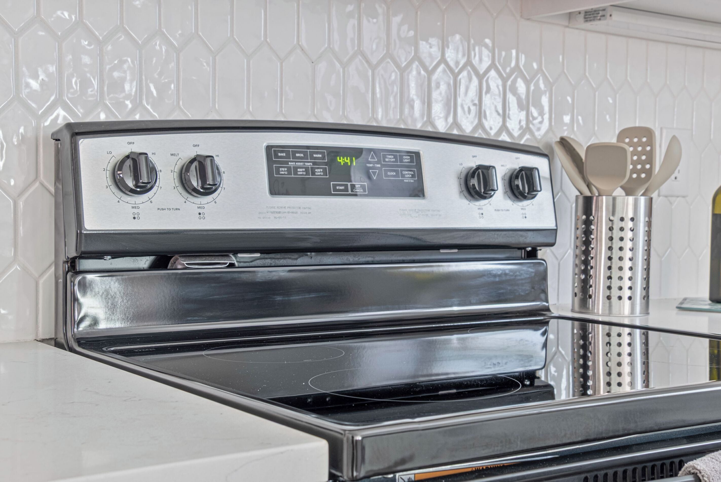 Une cuisine équipée d’une cuisinière en acier inoxydable, d’une horloge numérique affichant 4 h 41, et d’un porte-ustensiles contenant des spatules.