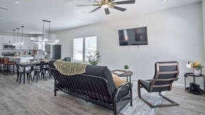 Ein modernes Wohnzimmer mit einer schwarzen Couch, einem Essbereich mit Tisch und Stühlen, einer Küche im Hintergrund und einem Deckenventilator.