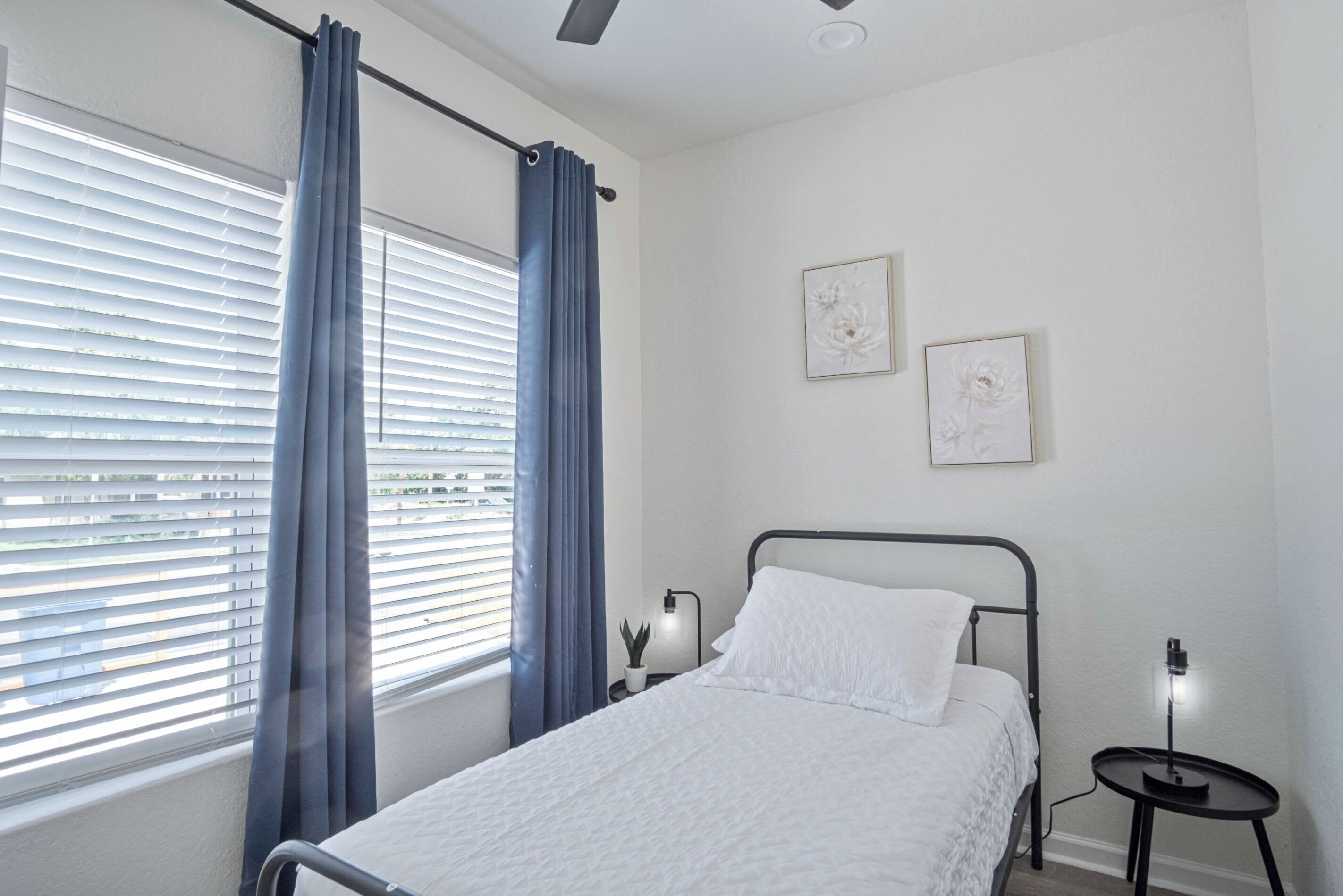 Un dormitorio con cama, una ventana con persianas, una mesita de noche con lámpara y dos cuadros colgados en la pared.