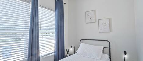 Un dormitorio con cama, una ventana con persianas, una mesita de noche con lámpara y dos cuadros colgados en la pared.