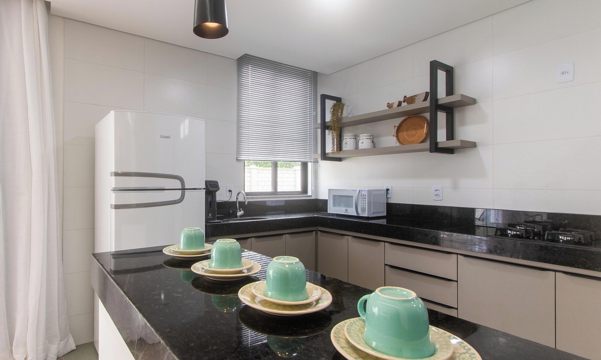 A modern kitchen with a refrigerator, microwave, and a sink. There are decorative items on shelves and a window with blinds.