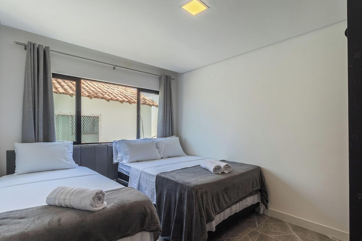 A hotel room with two beds, a window with curtains, a tiled floor, and a ceiling light.