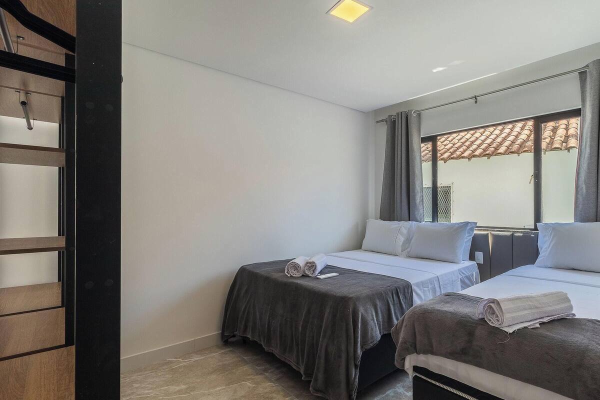 A hotel room with two beds, a window with curtains, a wooden shelf, and a ceiling light.