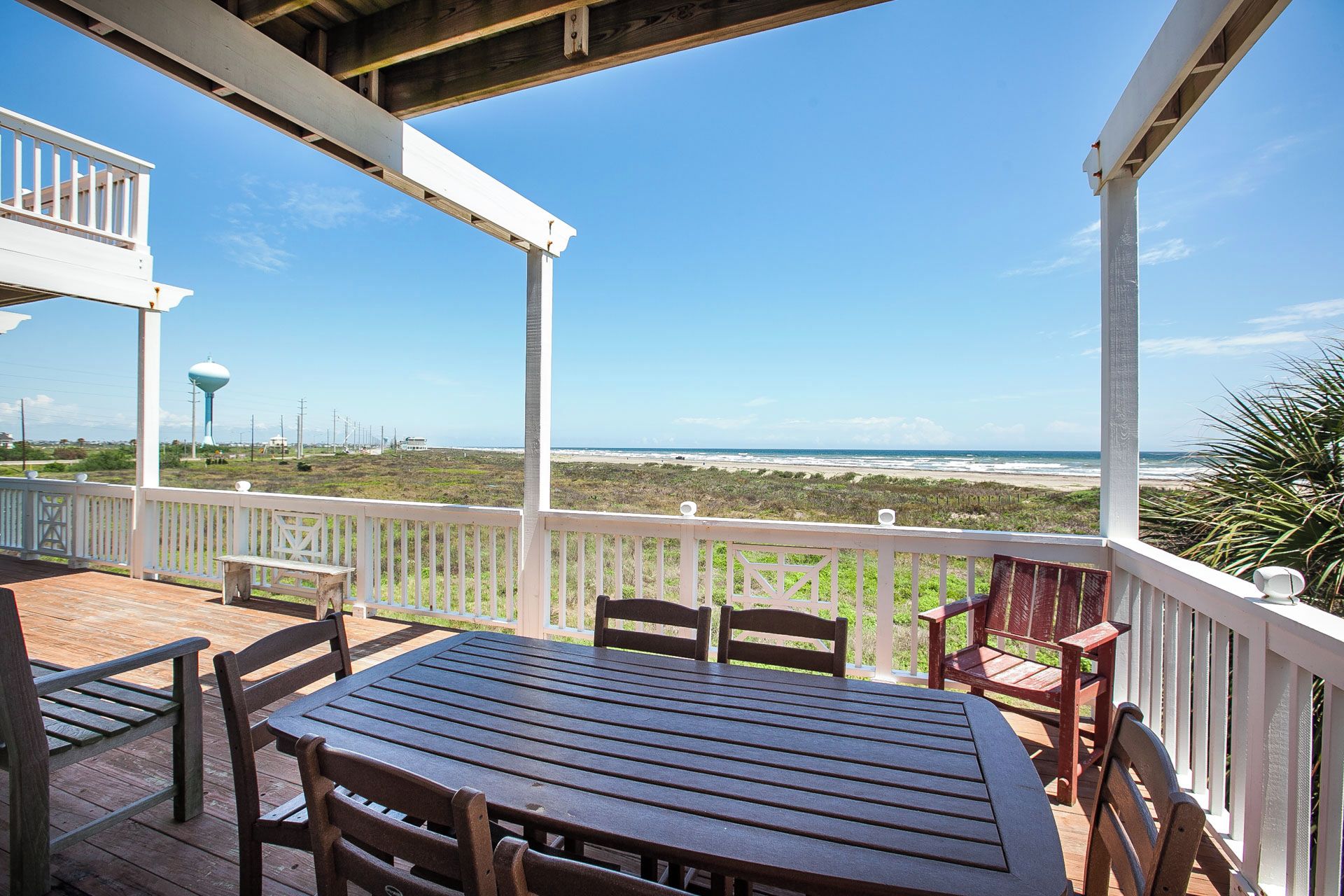 A wooden deck with a table and chairs, overlooking a beach with a water tower in the distance.