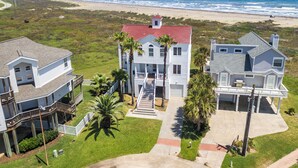 A coastal property with a two-story house featuring a red roof, a covered porch, and a front yard with palm trees. - Breakers Pointe by Ryson Vacation Rentals (Galveston)