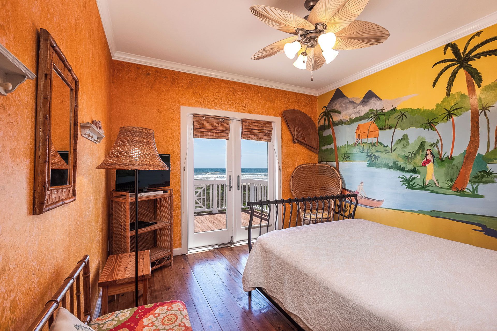 A room with a bed, a television on a wooden stand, wicker furniture, and a mural of a tropical scene.