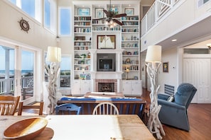 Ein geräumiges Wohnzimmer mit Kamin, einer großen Bücherwand und einem Fernseher. Es gibt einen Essbereich mit Tisch und Stühlen sowie einen Balkon mit Meerblick.