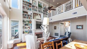 A spacious living area with a fireplace, bookshelves, a dining table, and a view of the outdoors. - Breakers Pointe by Ryson Vacation Rentals (Galveston)
