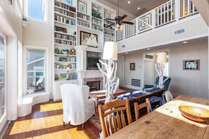 A spacious living area with a fireplace, bookshelves, a dining table, and a view of the outdoors. - Breakers Pointe by Ryson Vacation Rentals (Galveston)