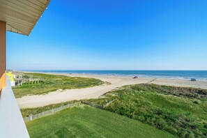 Vista a la playa desde un balcón con barandilla de madera, un camino de arena y una cerca de madera.