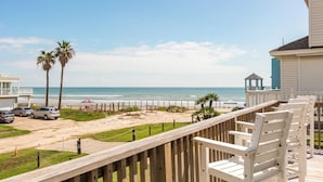 Eine Holzterrasse mit weißen Stühlen mit Blick auf einen Strand, Palmen und eine Laube.