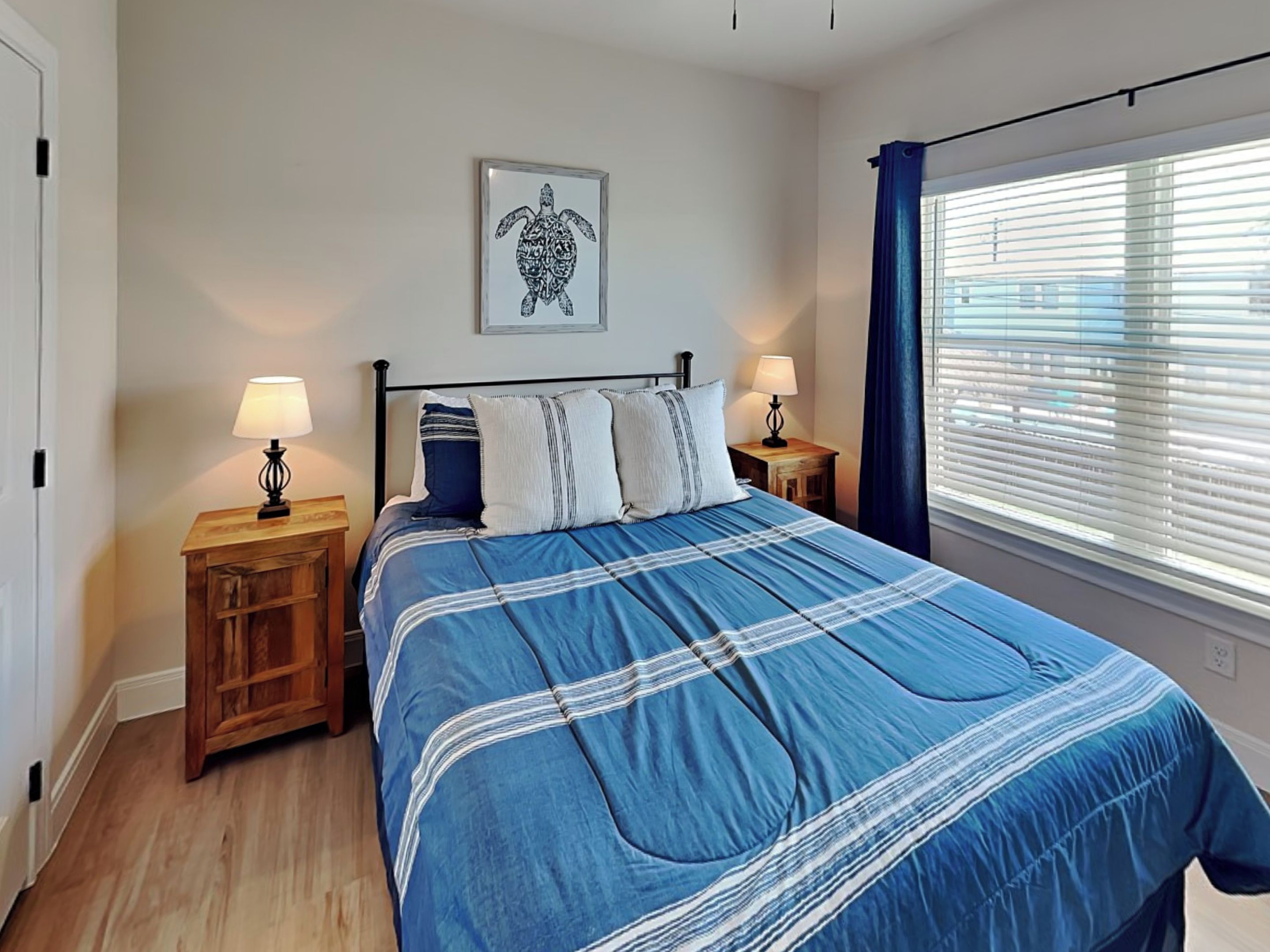 A bedroom with a bed, two bedside tables, a wall-mounted lamp, a window with blinds, and a framed picture on the wall.