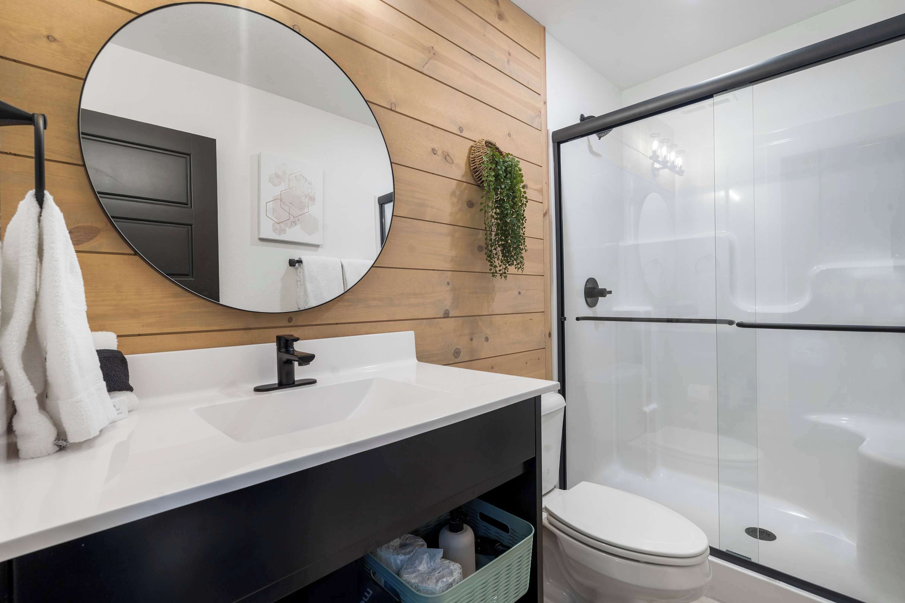 A modern bathroom with a wooden panel wall, a round mirror, a white sink, a black faucet, a white toilet, and a glass-enclosed shower.