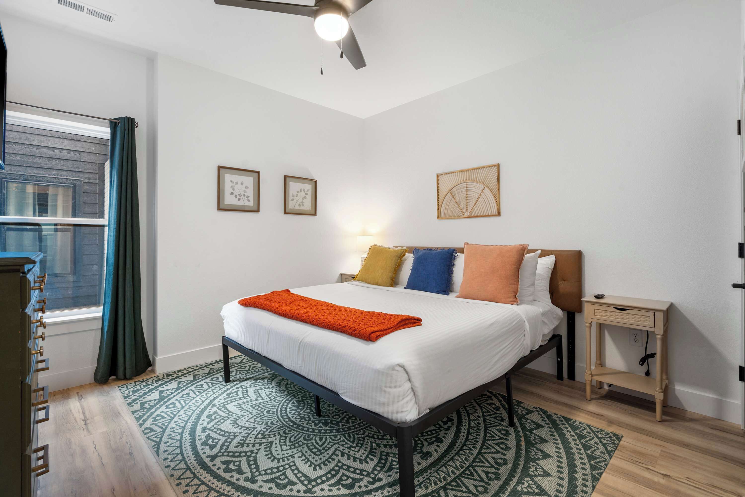 Une chambre à coucher bien rangée, avec un lit, une table de chevet, une fenêtre avec des rideaux et un tapis à motifs.