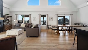 A spacious living area with a high ceiling, large windows, a dining table, and a sofa.