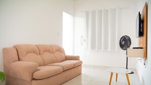 Un salon avec un canapé beige, une petite table blanche, un ventilateur sur pied et un téléviseur fixé au mur.